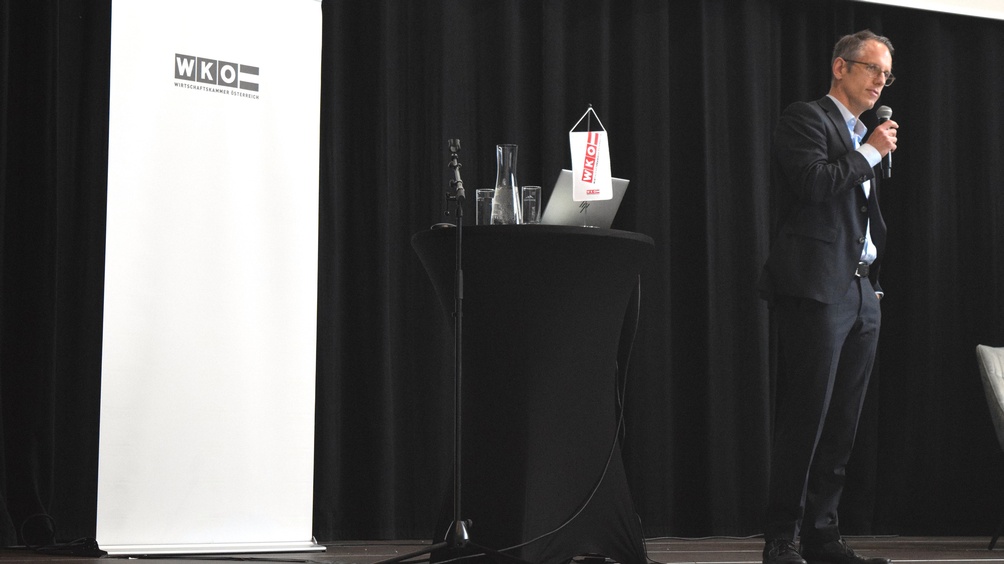 Eine Person steht neben einem Rundtisch, auf dem ein Laptop steht und ein Mikrofon liegt. Die Person spricht in ein Mikrofon. Hinter der Person ist ein weißer Banner, auf dem Wirtschaftskammer Österreich steht