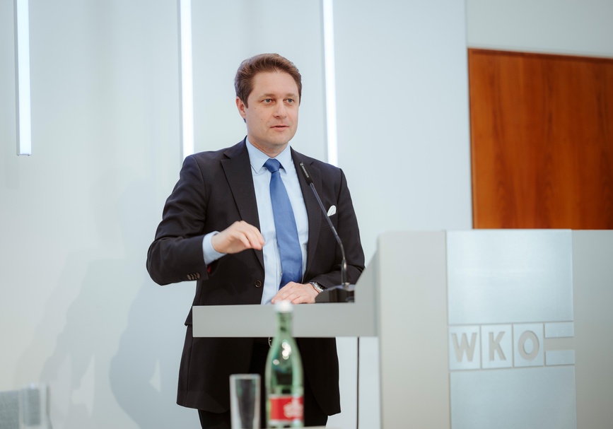 Frontale Aufnahme einer Person mit kurzen Haaren, Hemd und Sakko, die hinter einem Pult steht