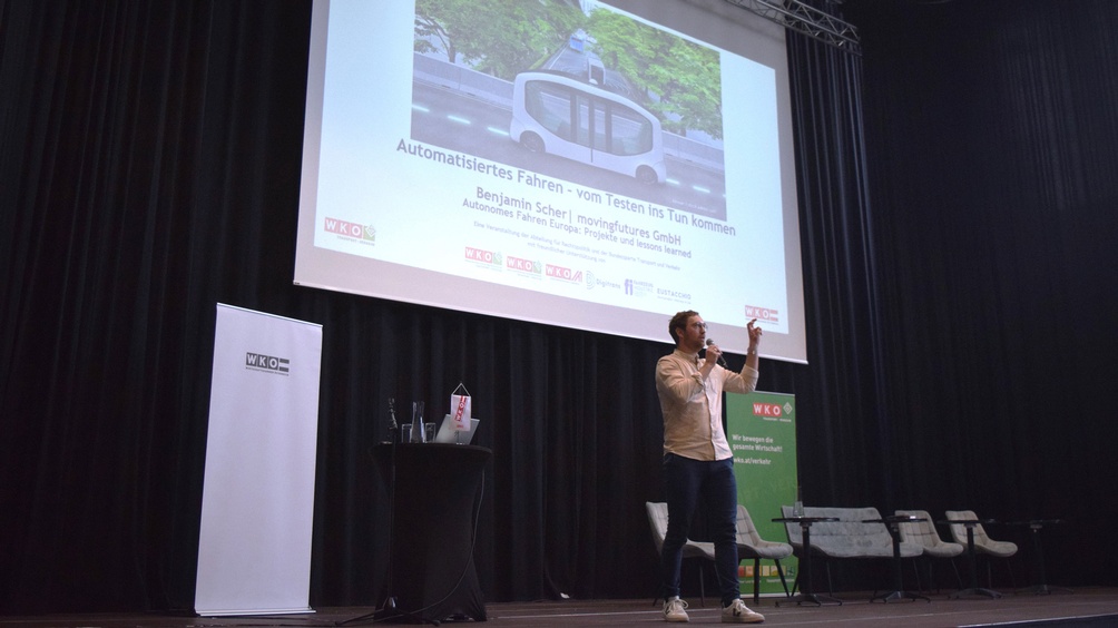 Eine Person steht neben einem Rundtisch, auf dem ein Laptop steht und ein Mikrofon liegt. Die Person spricht in ein Mikrofon. Hinter der Person ist ein weißer Banner, auf dem Wirtschaftskammer Österreich steht. Hinter der Person ist eine Leinwand, auf der automatisiertes Fahren - vom Testen ins Tun kommen steht