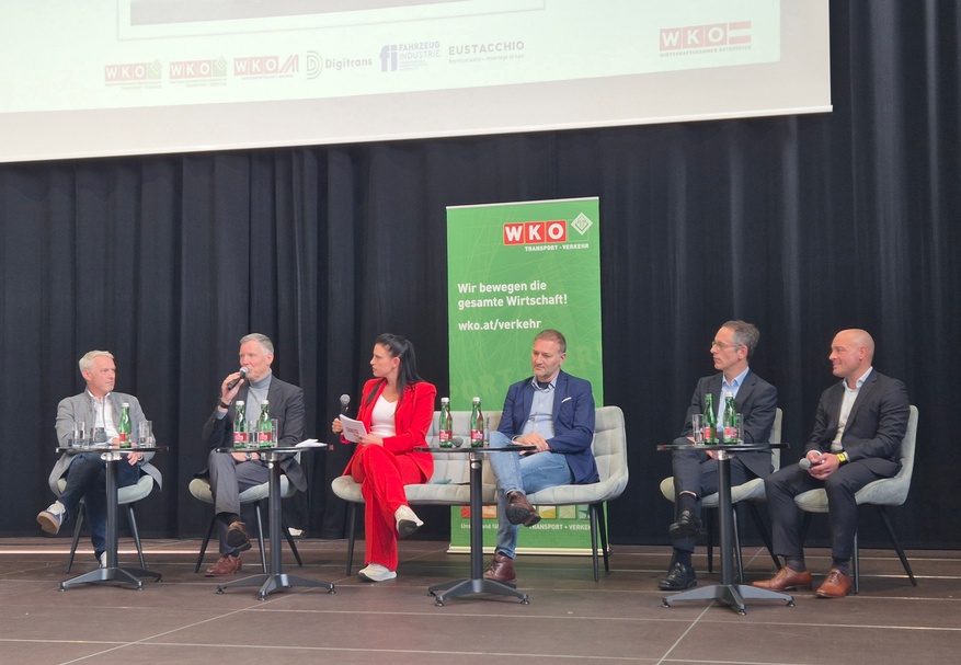 Auf einer Bühne sitzen nebeneinander auf sechs Stühlen sechs Personen. Eine der Personen hält ein Mikrofon und spricht in dieses. Hinter ihnen ist ein Banner, auf dem Wir bewegen die gesamte Wirtschaft steht