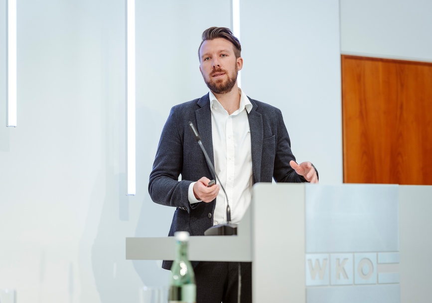 Frontale Aufnahme einer Person mit Bart, kurzen Haaren, Hemd und Sakko, die hinter einem Pult steht und in der rechten Hand eine kleine Fernbedienung hält