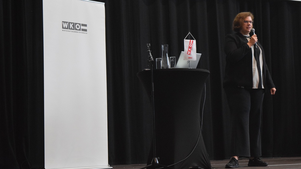 Eine Person steht neben einem Rundtisch, auf dem ein Laptop steht und ein Mikrofon liegt. Die Person spricht in ein Mikrofon. Hinter der Person ist ein weißer Banner, auf dem Wirtschaftskammer Österreich steht