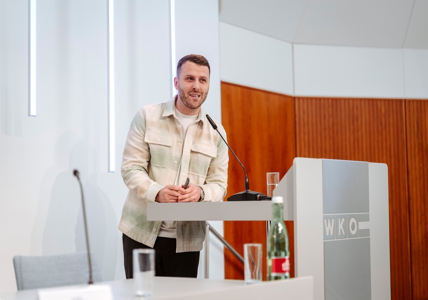 Frontale Aufnahme einer Person mit kurzen Haaren, Bart und kleiner Fernbedienung in den Händen, die hinter einem Pult steht und in ein Mikrofon spricht