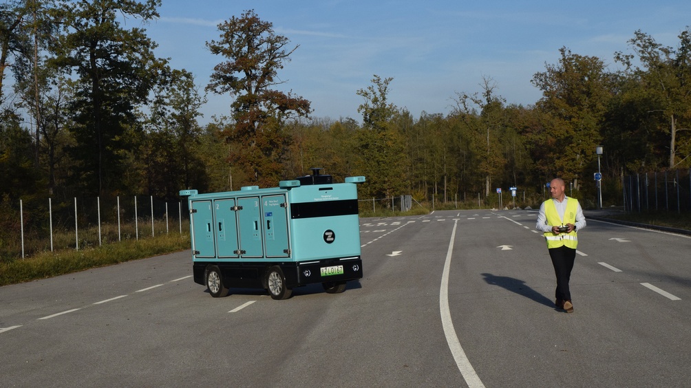 Auf einer mehrspurigen Straße steht ein automatisch fahrendes Fahrzeug mit vier Fächern und vier Rädern. Neben dem Fahrzeug ist eine Person, die das Fahrzeug mit einer Fernbedienung steuert