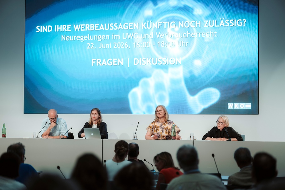 Vier Personen sitzen an einem Podium vor einer großen Projektionsfläche. Im Vordergrund sind Teilnehmende der Veranstaltung zu sehen. Auf der Projektionsfläche steht: „SIND IHRE WERBEAUSSAGEN KÜNFTIG NOCH ZULÄSSIG?<br />
Neuregelungen im UWG und Verbraucherrecht, FRAGEN | DISKUSSION“