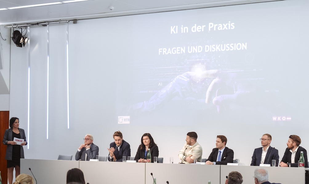 Frontale Aufnahme von sieben Personen, die an der Längsseite eines Tisches sitzen. Vor den Personen sind Mikrofone, Laptops und Wassergläser. Hinter ihnen ist eine große Leinwand, auf der KI in der Praxis. Ihnen gegenüber sitzt ein Publikum