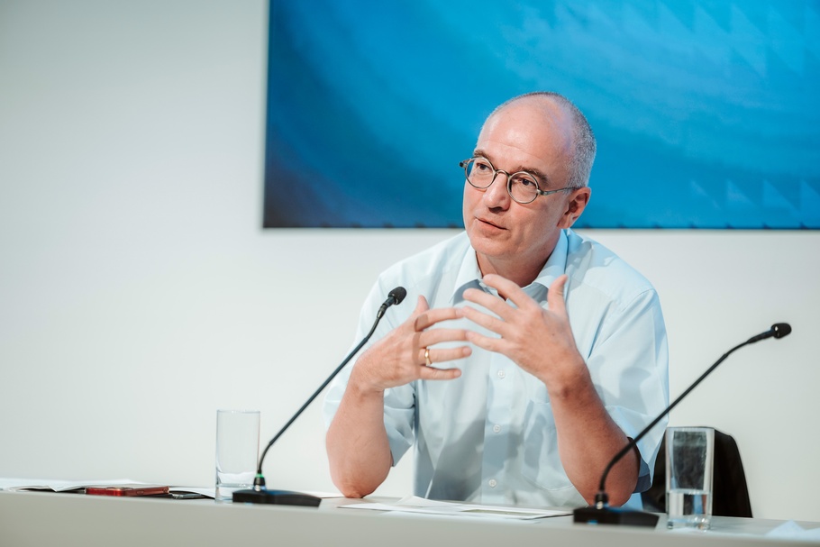 Eine Person sitzt an einem Podiumstisch und spricht. Vor der Person stehen zwei Tischmikrofone. Auf dem Tisch liegen Unterlagen sowie zwei Gläser mit Wasser. Im Hintergrund ist ein blauer Bildschirm oder eine Projektionsfläche