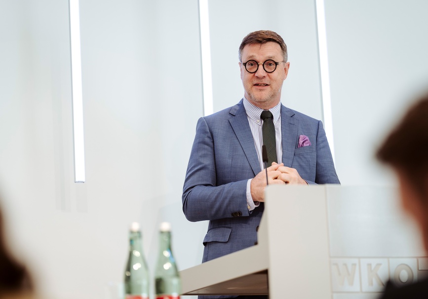 Frontale Aufnahme einer Person mit kurzen Haaren, Bart und Brille, die hinter einem Pult steht und in ein Mikrofon spricht