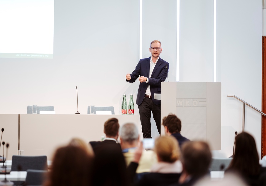 Frontale Aufnahme einer Person in einem blauen Oberteil, mit Sakko und Brillen, die hinter einem Pult in einem Raum steht und in Mikrofon spricht. Ihr gegenüber sitzt Personen in einem Publikum