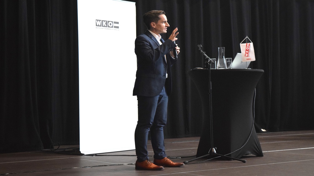 Eine Person steht neben einem Rundtisch, auf dem ein Laptop steht und ein Mikrofon liegt. Die Person spricht in ein Mikrofon. Hinter der Person ist ein weißer Banner, auf dem Wirtschaftskammer Österreich steht