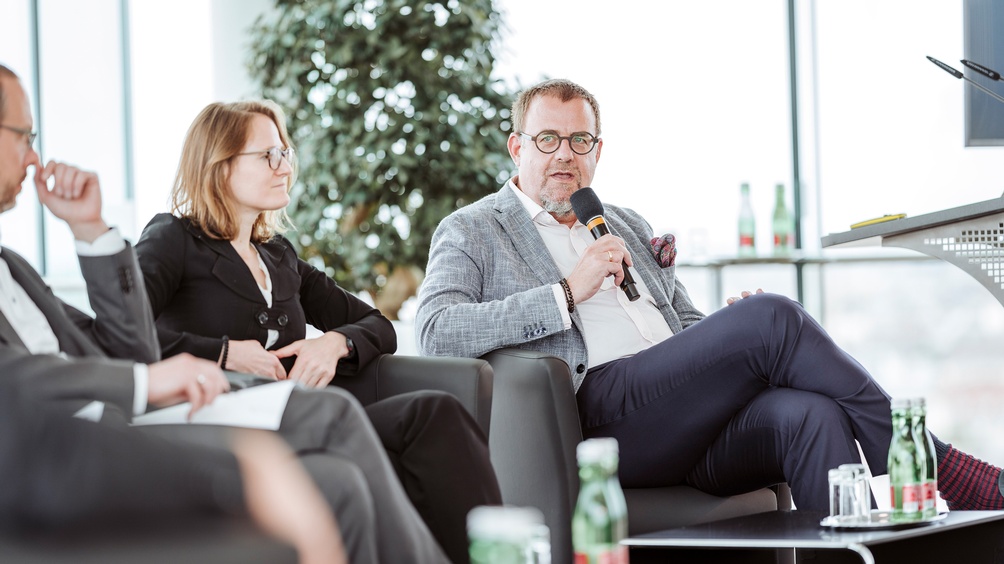 Drei sitzende Personen, eine Person spricht in Mikrofon