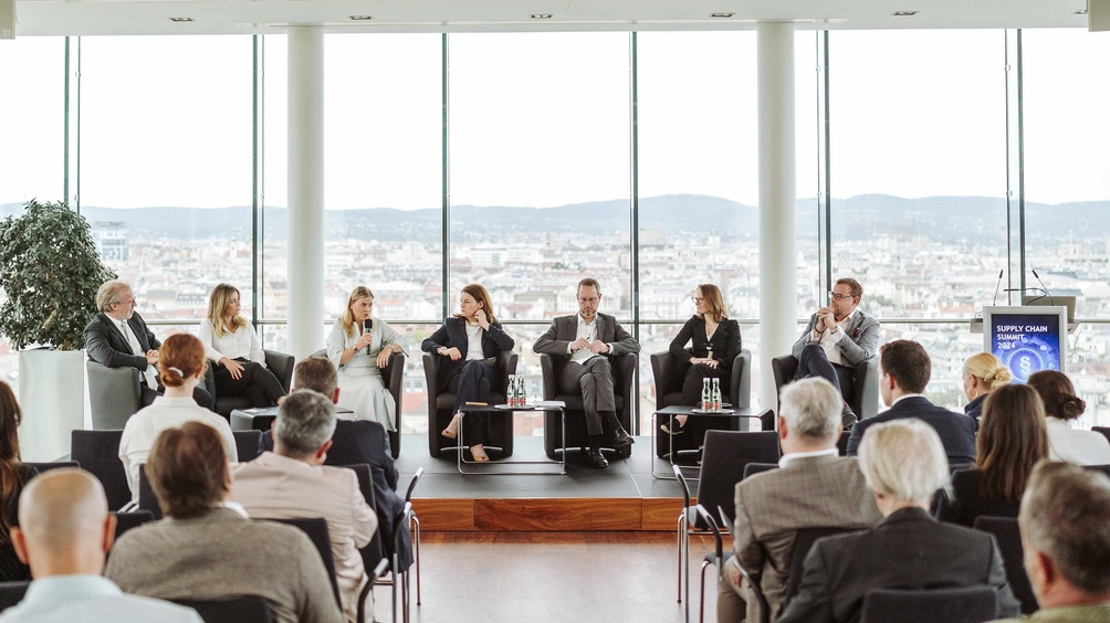 Mehrere Personen sitzen auf einer Bühne vor einem Publikum, eine Person spricht in ein Mikrofon, im Hintergrund Blick auf eine Stadtlandschaft