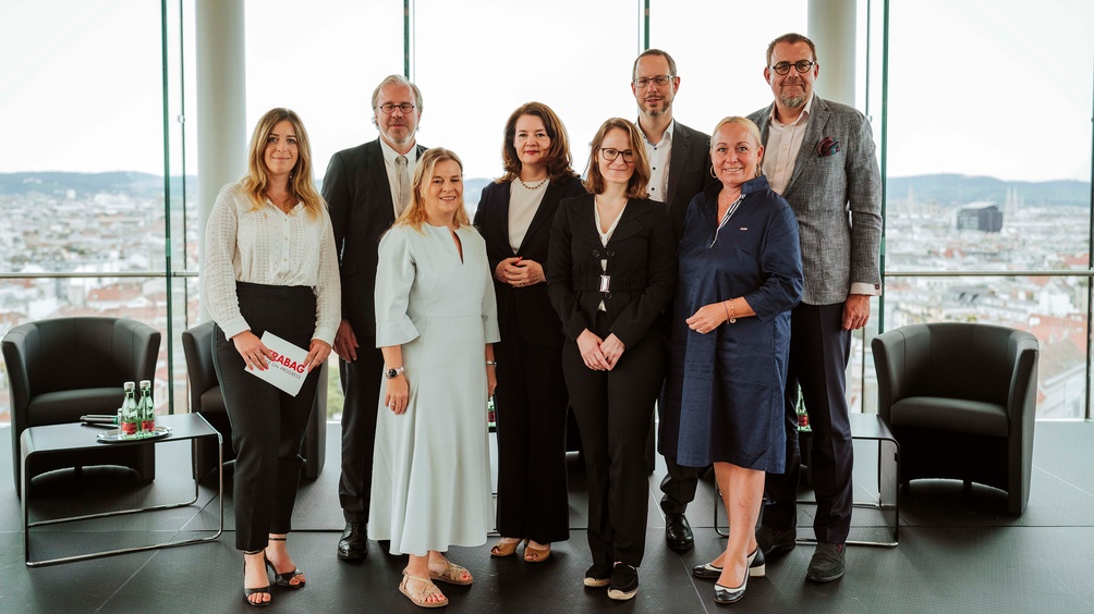 Mehrere Personen neben- und hintereinander stehend vor großem Glasfenster, das Blick auf Stadtlandschaft bietet