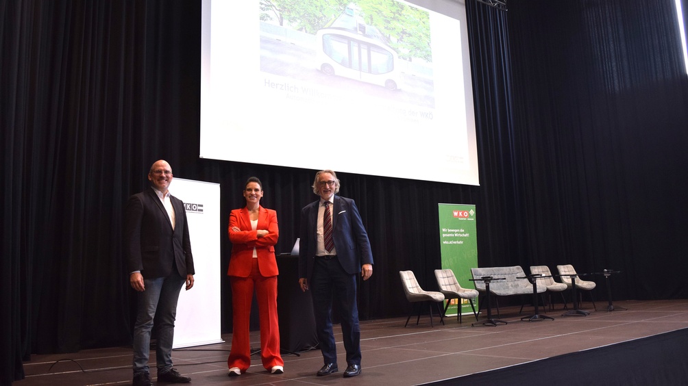 Porträt von drei Personen in Sakkos, die nebeneinander auf einer Bühne stehen. Hinter ihnen ist ein weißer Banner, ein Stehtisch, eine große Leinwand, ein paar Sesseln und ein weiterer Banner, auf dem wir bewegen die gesamte Wirtschaft steht