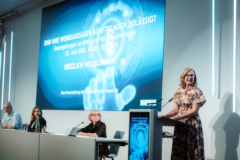Eine Person steht an einem Rednerpult und spricht vor Publikum. Links im Bild sitzen drei weitere Personen an einem Podiumstisch. Auf dem Rednerpult ist ein Bildschirm mit dem Veranstaltungsdesign. Auf der Projektionsfläche im Hintergrund steht „SIND IHRE WERBEAUSSAGEN KÜNFTIG NOCH ZULÄSSIG? Neuregelungen im UWG und Verbraucherrecht. HERZLICH WILLKOMMEN! Eine Veranstaltung der Abteilung für Rechtspolitik