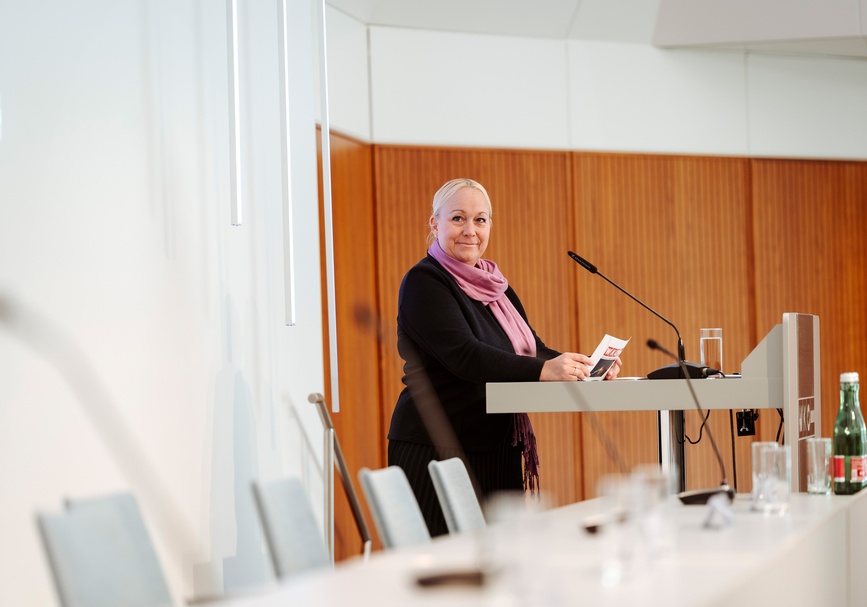  Frontale Aufnahme einer Person mit hellen, geschlossenen Haaren, schwarzem Oberteil und rosa Schal, die hinter einem Pult steht und in ein Mikrofon spricht