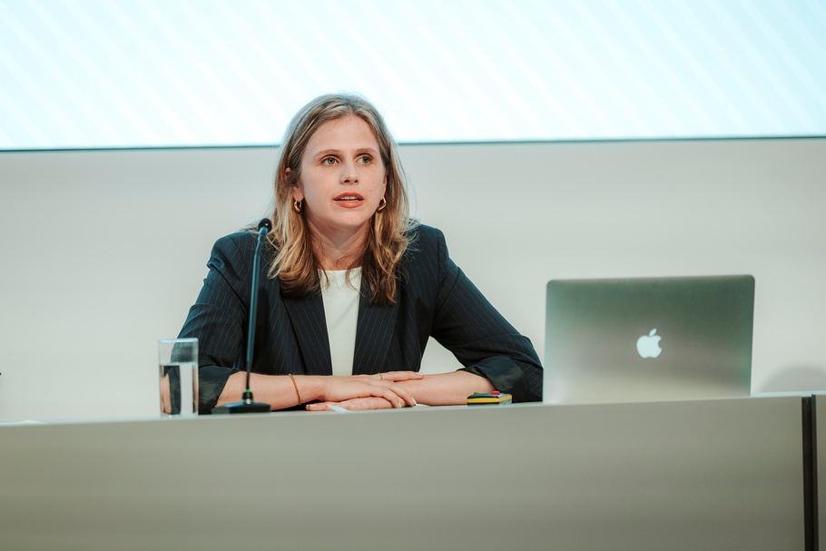 Eine Person sitzt an einem Podiumstisch. Vor der Person stehen ein Mikrofon, ein Laptop und ein Wasserglas