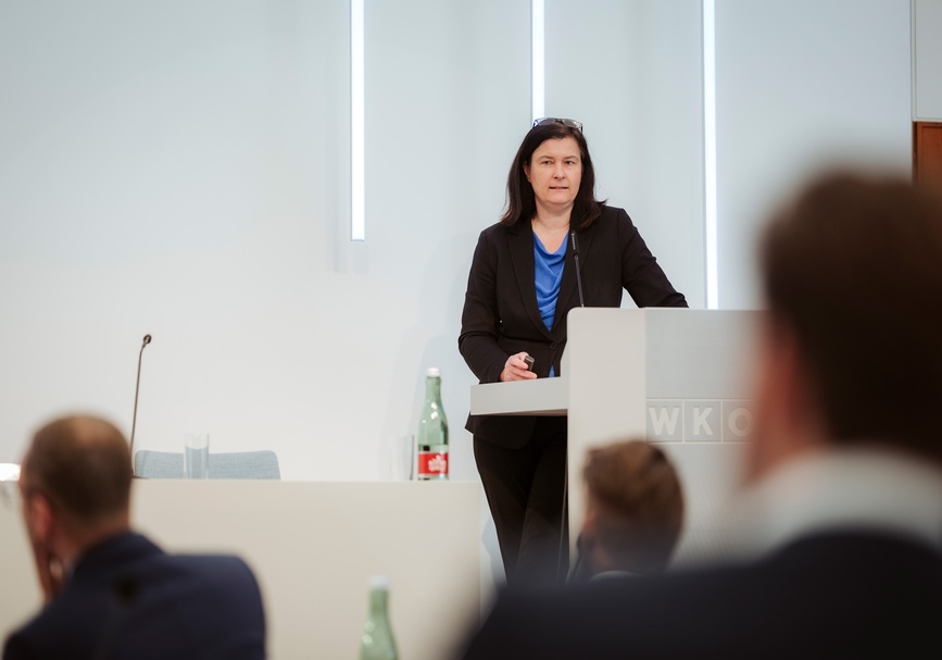 Frontale Aufnahme einer Person in einem blauen Oberteil, mit Sakko und Brillen in den Haaren, die hinter einem Pult in einem Raum steht und in Mikrofon spricht. Ihr gegenüber sitzt Personen in einem Publikum