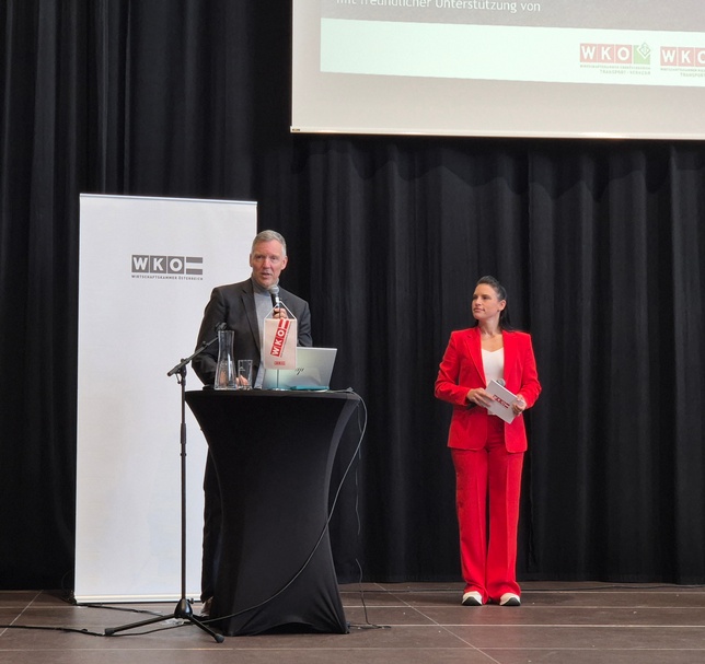 Eine Person steht hinter einem Pult auf einer Bühne und spricht in ein Mikrofon. Rechts neben der Person steht eine weitere Person, die Moderationskarten hält. Hinter der Person hängt eine große Leinwand