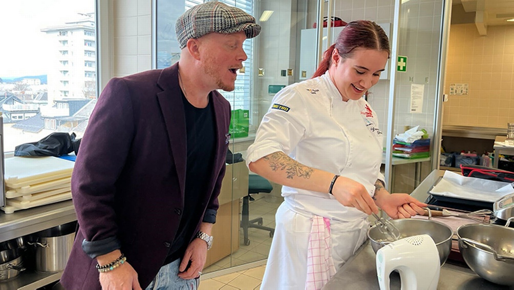 Person mit Kappe schaut in Küche anderer Person beim Kochen über die Schulter