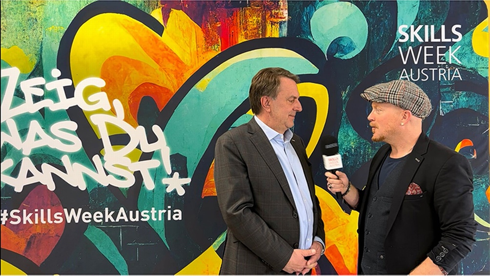 Zwei Personen in Interviewsituation vor Grafiti, eine Person hält Mikrofon