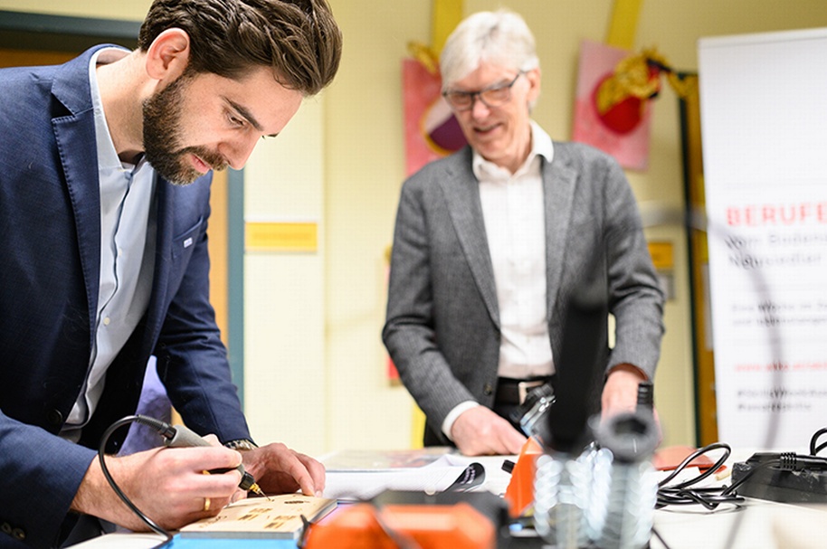 Eine Person über Tisch gebeugt mit Gerät Brett bearbeitend, eine weitere Person beobachtend