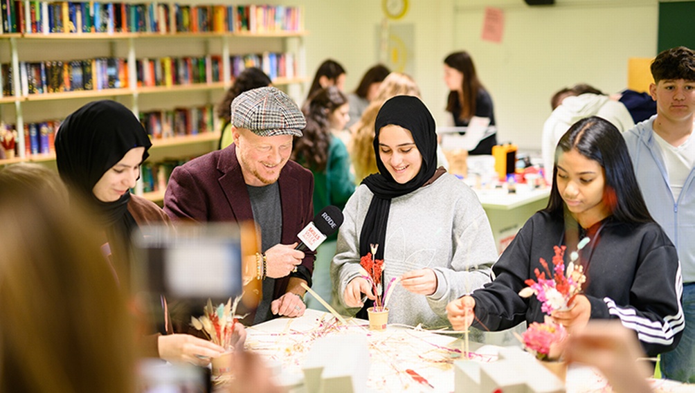Mehrere Kinder auf Tisch blickend, weitere Person mit Mikrofon interviewt eines der Kinder