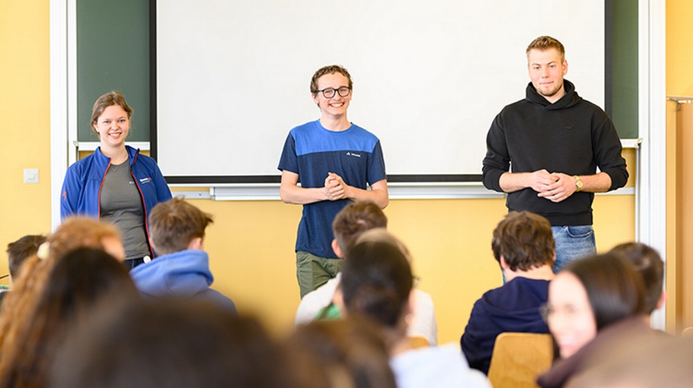 Drei Personen Tafel mit Leinwand stehend ins Publikum blickend