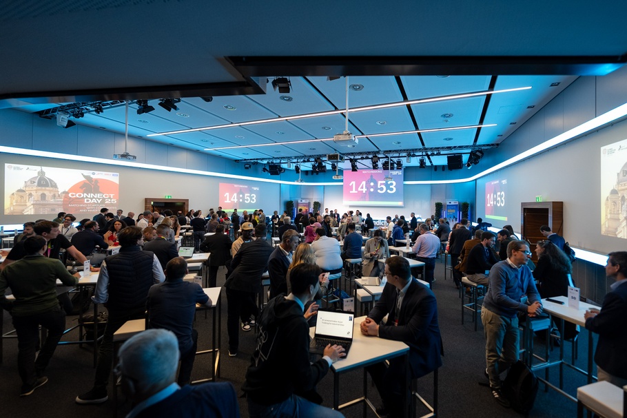 Veranstaltung mit vielen Menschen, die an kleinen Tischen vor ihren Laptops sitzen. Sie befinden sich in einem Veranstaltungssaal, auf der hinteren Wand des Saals ist ein Fernseher montiert, auf dem ein Timer abläuft
