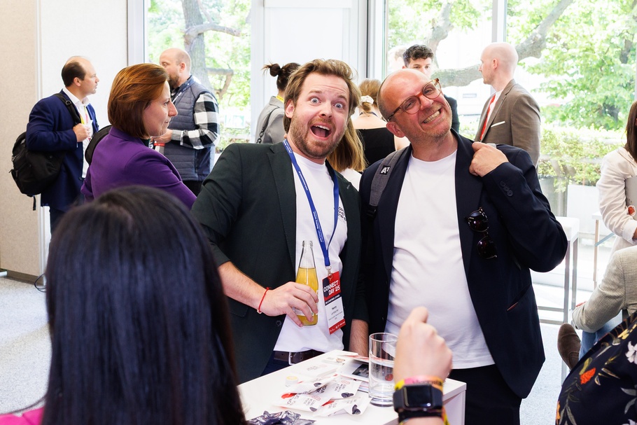 Nahaufnahme von zwei gut gelaunten Menschen während einer Veranstaltung. Sie haben die Hände auf den Rücken des anderen gelegt, die rechte Person hält eine Glasflasche mit gelber Limonade in der Hand. , im Hintergrund sind andere Personen zu sehen