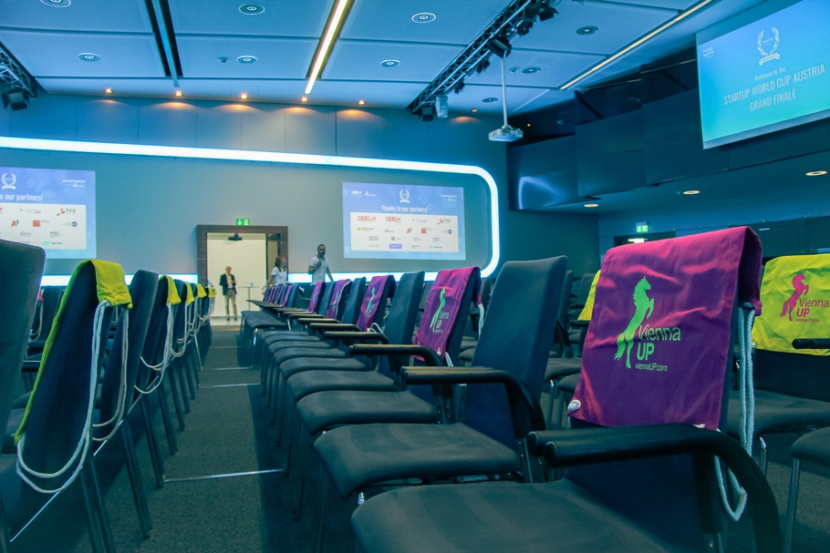 Nahaufnahme eines Veranstaltungssaals, in dem Stühle nebeneinander in Reihen platziert sind. Über den Stühlen hängen bunte Jutebeutel mit dem Schriftzug ViennaUP