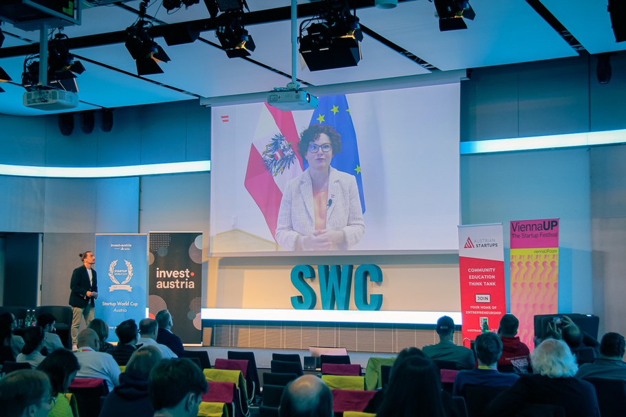 Aufnahme eines Veranstaltungsaals, in dem Stuhlreihen sind, auf den Stühlen sitzen Personen. Sie schauen auf einen Bildschirm, der über einer Bühne platziert ist 