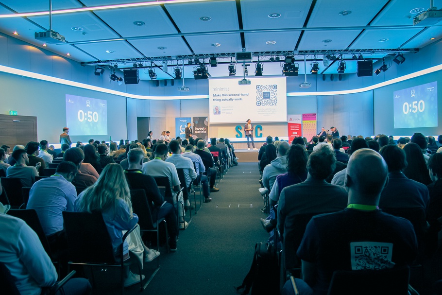 Aufnahme eines Veranstaltungsraums, in dem Personen nebeneinander in mehreren Reihen sitzen. Sie schauen auf einen Bildschirm, der über einer Bühne platziert ist. Rechts neben der Bühne ist eine Person, die diese betreten möchte