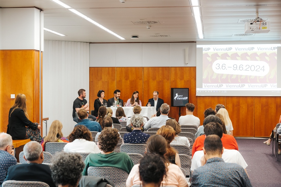 Vortragssaal mit Publikum und fünf Sprecher:innen an einem Tisch 