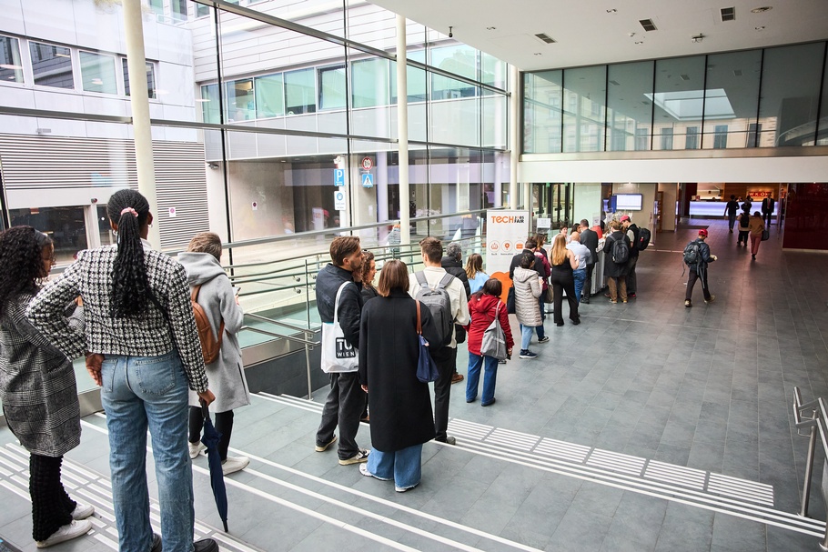 Menschen stehen in einer Reihe nebeneinander und sind von hinten fotografiert. Sie warten im Foyer auf den Check-In einer Veranstaltung 