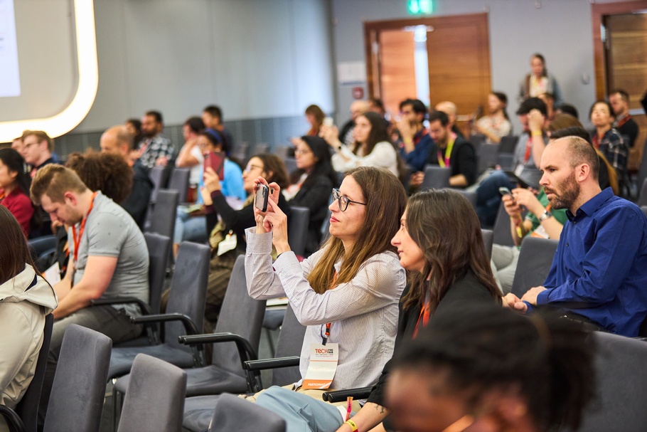 Personen sitzen nebeneinander auf Sesseln, die in Reihen aufgestellt sind. Eine Person macht ein Selfie mit der Kamera ihres Smartphones 