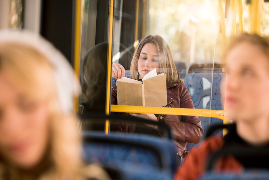 Eine Person sitzt in einem Bus auf einem Sitz und liest in einem Buch. Im Vordergrund verschwommen sind weitere Personen.