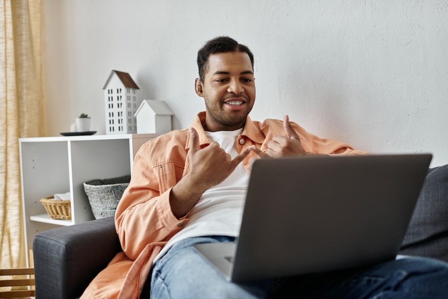 Eine Person mit Zahnspange, kurzen, dunklen Haaren und Hörgerät sitzt auf einem Sofa und gestikuliert mit beiden Händen. Die Person hält die Hände vor ihren Oberkörper. Auf dem Schoß hat sie einen aufgeklappten Laptop