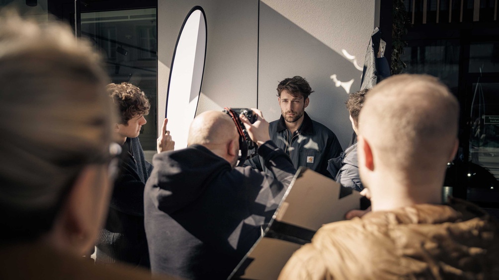 Szene aus einem Fotoshooting, mehrere Personen stehen um einen Mann herum, der gerade in die Kamera lächelt