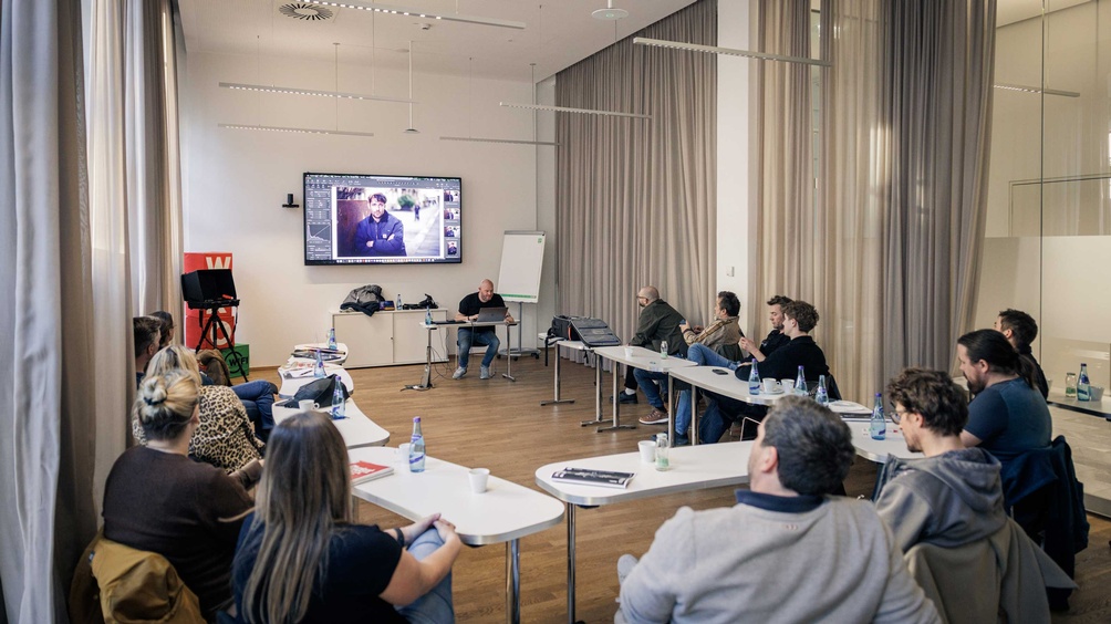 Mehrere Personen sitzen in einem Seminarraum. Vorne sitzt ein Referent und man sieht ein Foto auf einem großen Fernseher.