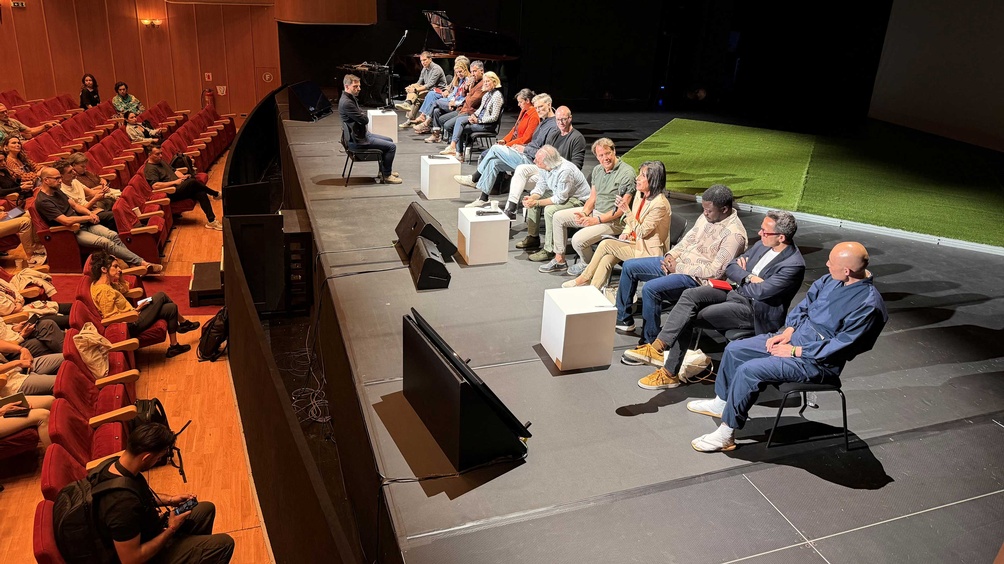 Rechts im Bild sieht man eine Bühne, auf der 14 Menschen in einer Reihe nebeneinander auf Stühlen sitzen. Eine Dame aus dieser Reihe spricht in ein Mikrofon. Links im Bild sieht man Menschen im Publikum sitzen.