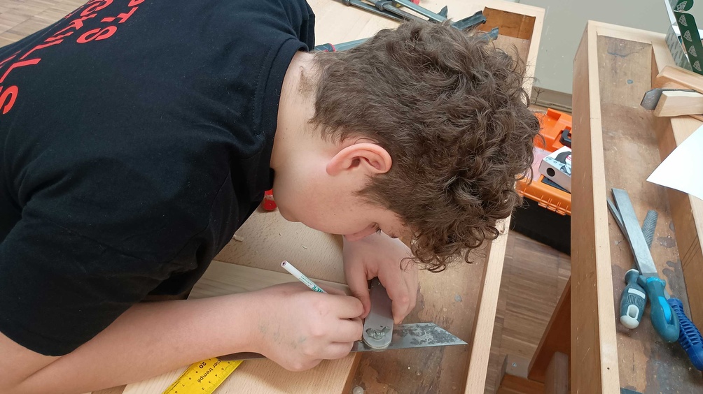 Ein Jugendlicher arbeitet an einem Werkstück aus Holz und zeichnet mti einem Bleistift etwas ein
