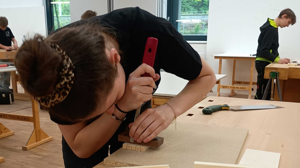Einge Jugendliche arbeitet mit einem Stemmeisen an einem Stück Holz, im Hintergrund sieht man mehrere andere Jugendliche an ihren Werkbänken