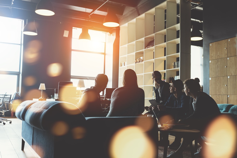 Gruppe von Personen sitzt rund um einen Tisch in einem Büro und hält eine Besprechung. Die Sonne scheint so durch das Fenster, dass nur die Silhouetten der Personen zu erkennen sind.