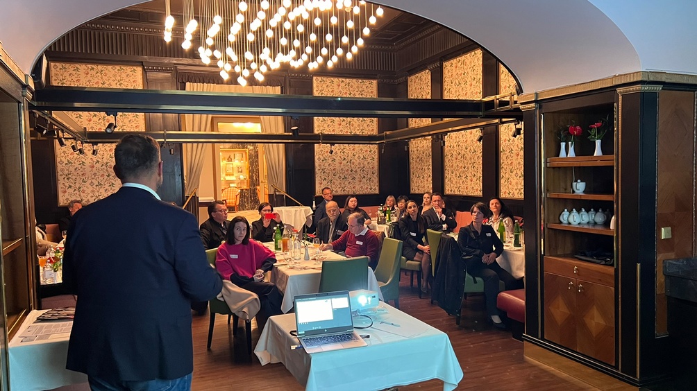 Person in Rückenansicht vor Laptop und auf grünen Stühlen an Tischen sitzendem Publikum sprechend