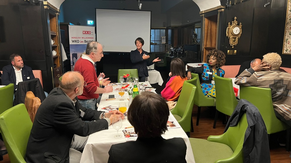 Person vor großer Videowall stehend und vor auf grünen Stühlen an Tischen sitzendem Publikum sprechend