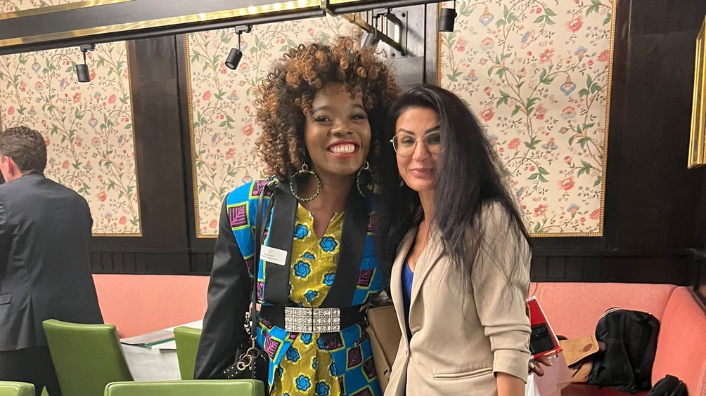 Zwei lächelnde Personen eng beieinander stehend vor Wand mit floraler Musterung und Holzvertäfelung