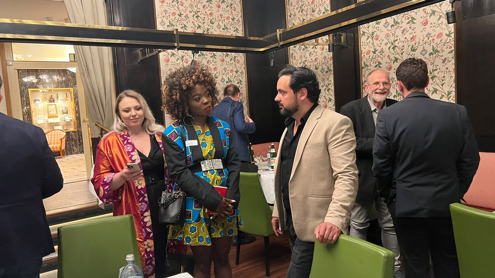 Zwei Personen im Gespräch miteinander, eine Person stützt Hände an grünem Stuhl ab, im Hintergrund floral gemusterte Tapete, Holzvertäfelung und weitere Personen im Gespräch