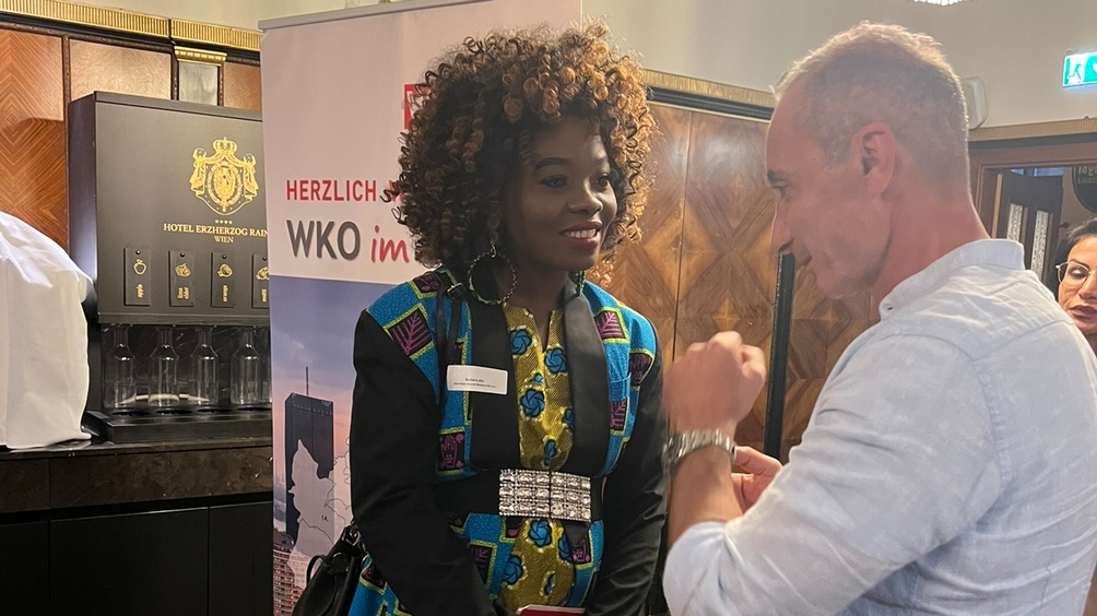 Zwei lächelnde Personen im Gespräch miteinander, im Hintergrund Rollup mit Schriftzug Herzlich WKO im und holzvertäfelte Wand