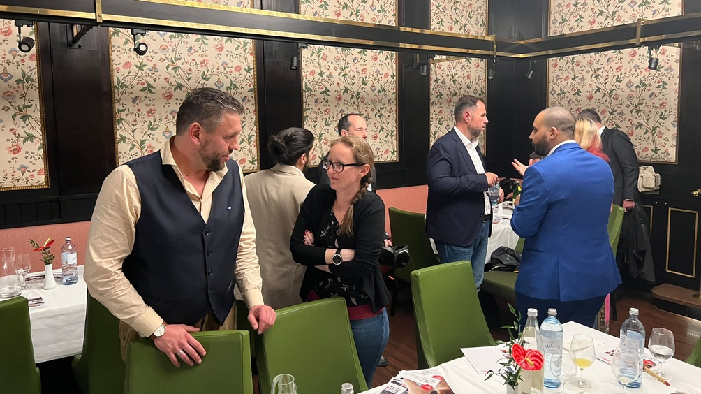 Zwei Personen im Gespräch miteinander, eine Person stützt Hände an grünem Stuhl ab, im Hintergrund floral gemusterte Tapete, Holzvertäfelung und weitere Personen im Gespräch